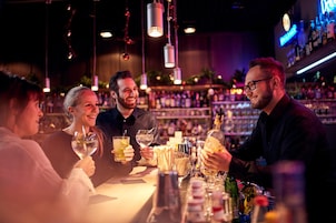 Um bar com pessoas sentadas em mesas, segurando bebidas, e um barman atrás do balcão.
