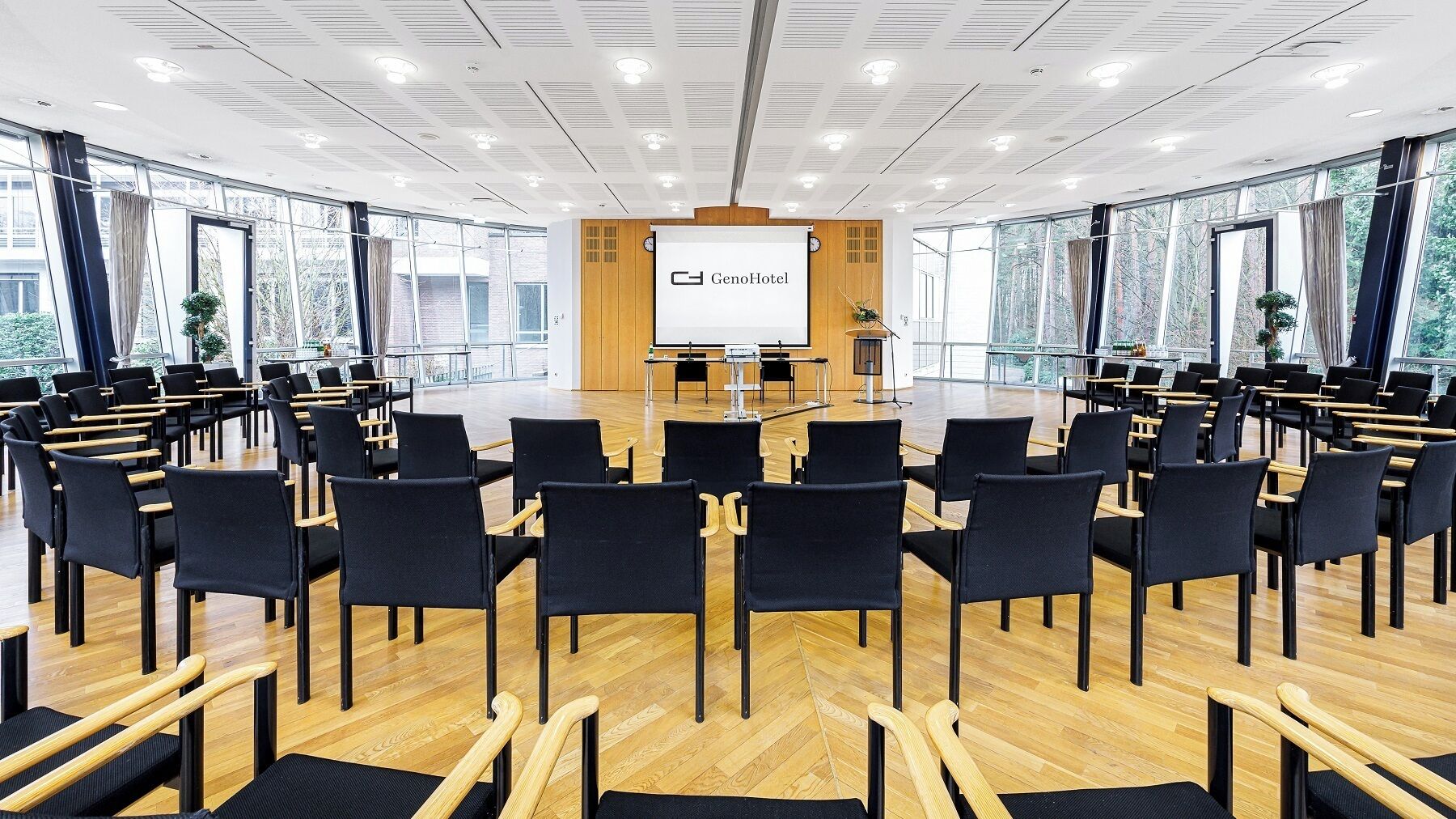 Un salón de conferencias con una pantalla grande al frente, filas de sillas y ventanas amplias.