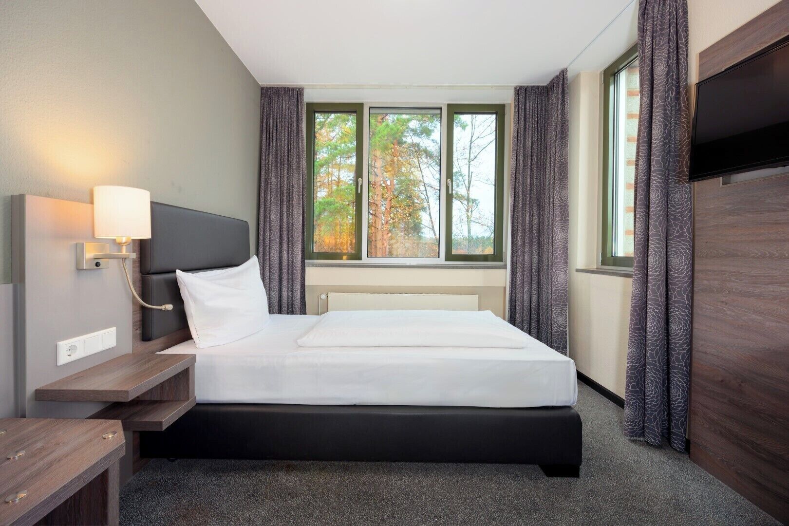 A hotel room with a bed, a bedside table, a lamp, a TV, and a window with curtains.