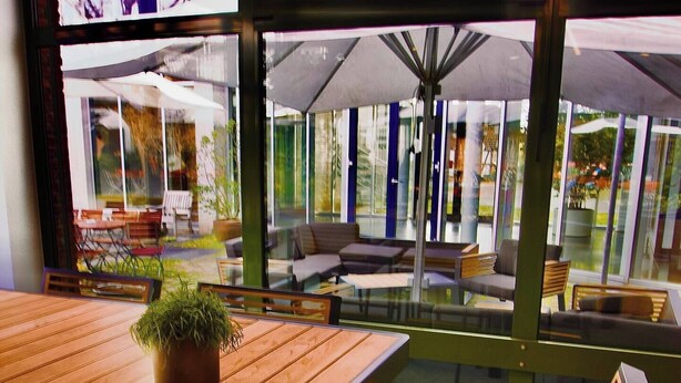 An indoor seating area with wooden tables and benches, large windows, and a view of the outdoor seating area. - GenoHotel Forsbach (Roesrath)