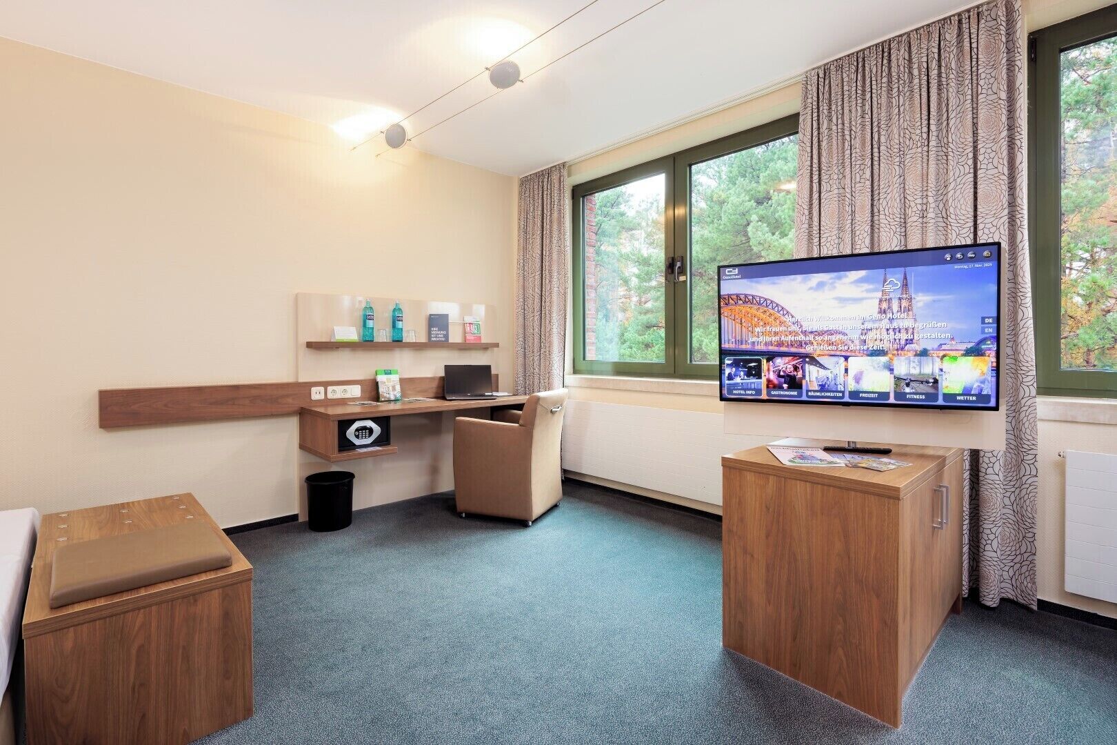 Quarto de hotel com escrivaninha, cadeira, televisão e janela com cortinas.