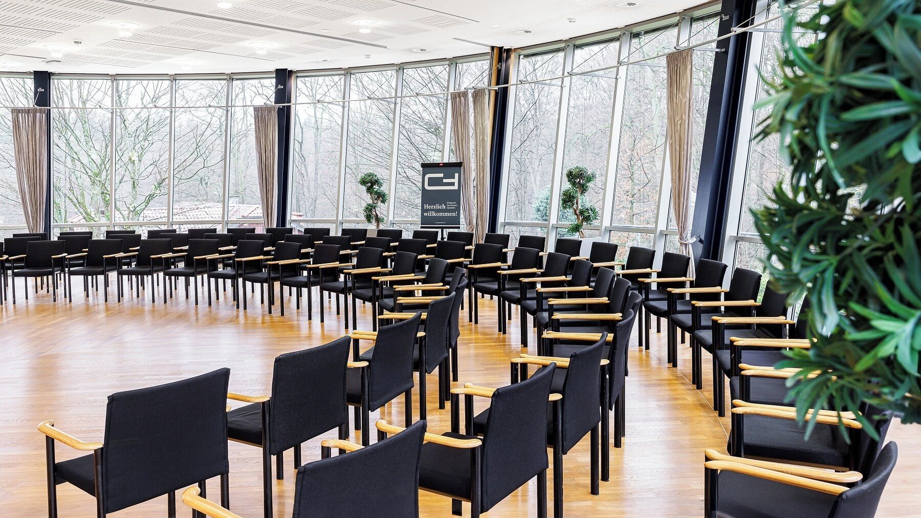 Un salón de conferencias con filas de sillas negras y mesas de madera, ventanas grandes con cortinas y un cartel con texto.