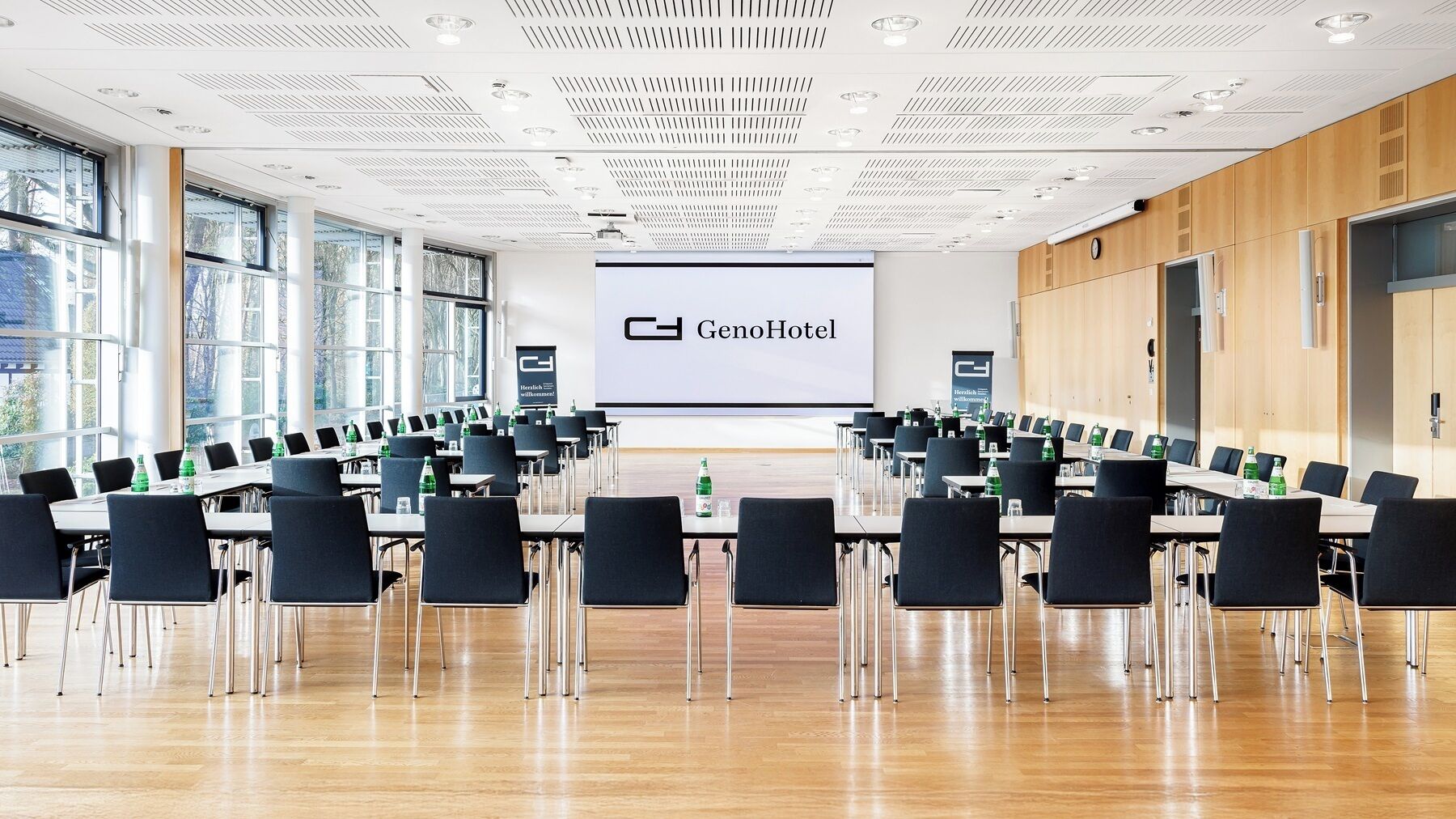 A conference room with a large screen displaying "GenoHotel" and a logo, arranged with tables and chairs, large windows, and a wooden wall.