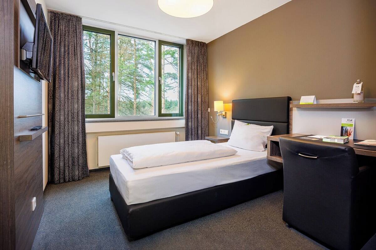 Habitación de hotel con cama, escritorio, silla, televisor, lámpara y ventana con cortinas.