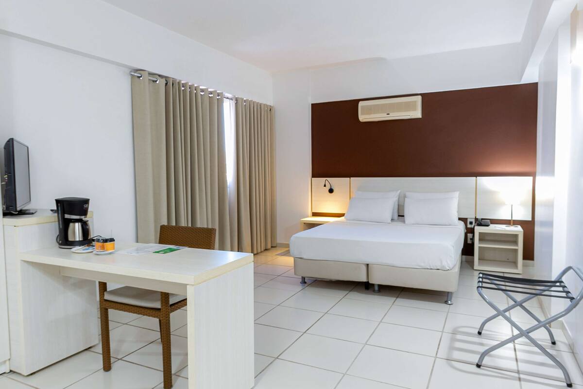 Habitación de hotel con cama, escritorio, silla, televisión, cafetera y ventana con cortinas.