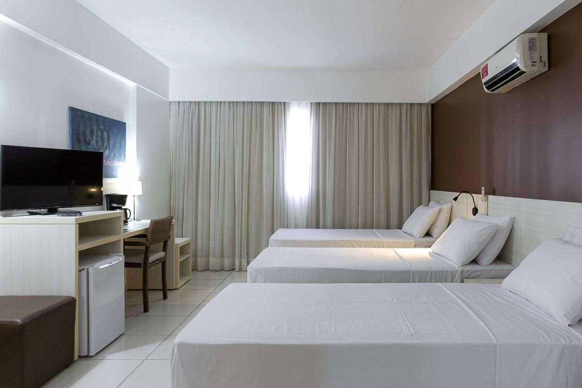 Habitación de hotel con dos camas, un escritorio con televisor, una silla, una mesita y un equipo de aire acondicionado.