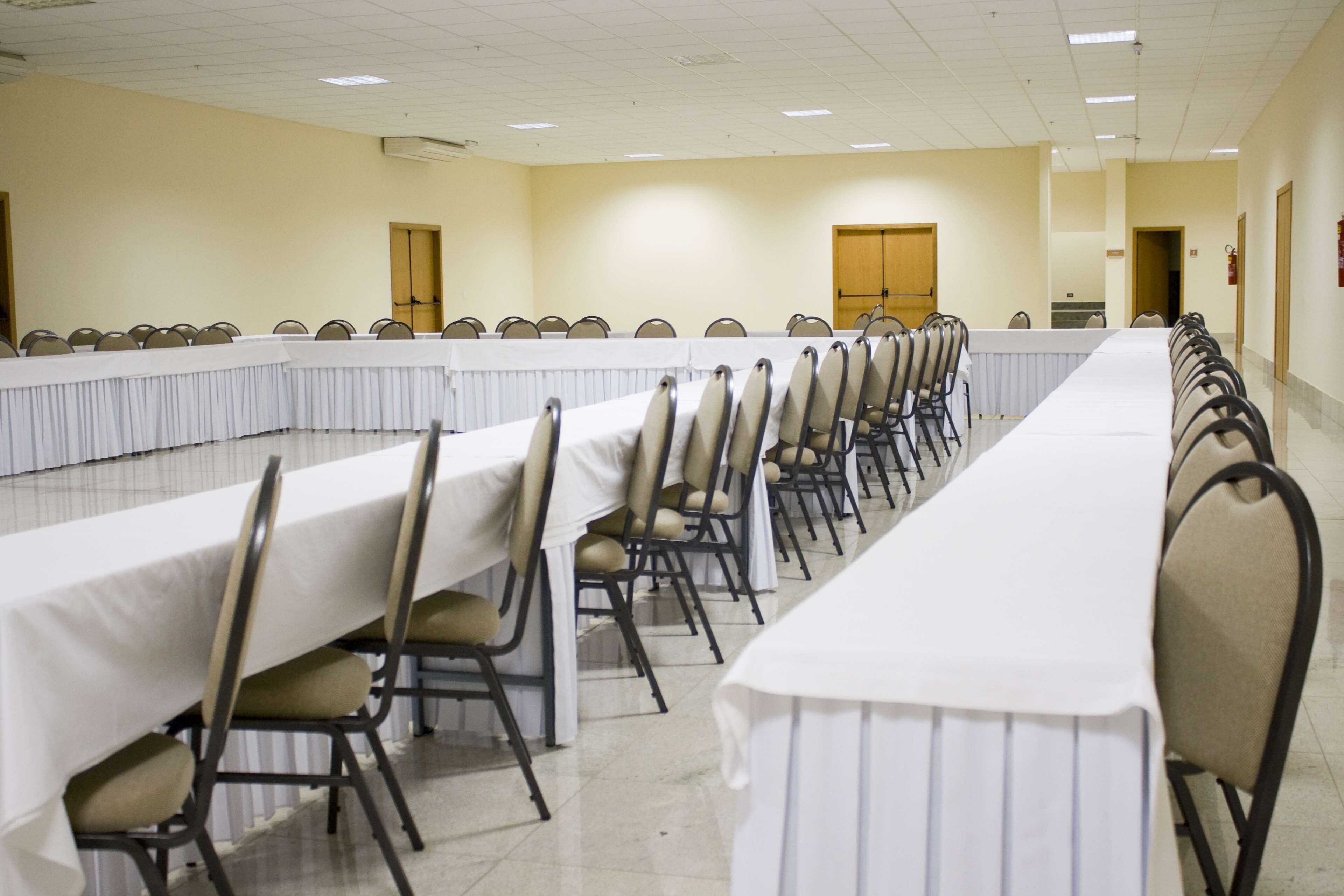 A conference room with long tables and chairs, beige cushioned chairs, white tablecloths, and a tiled floor.
