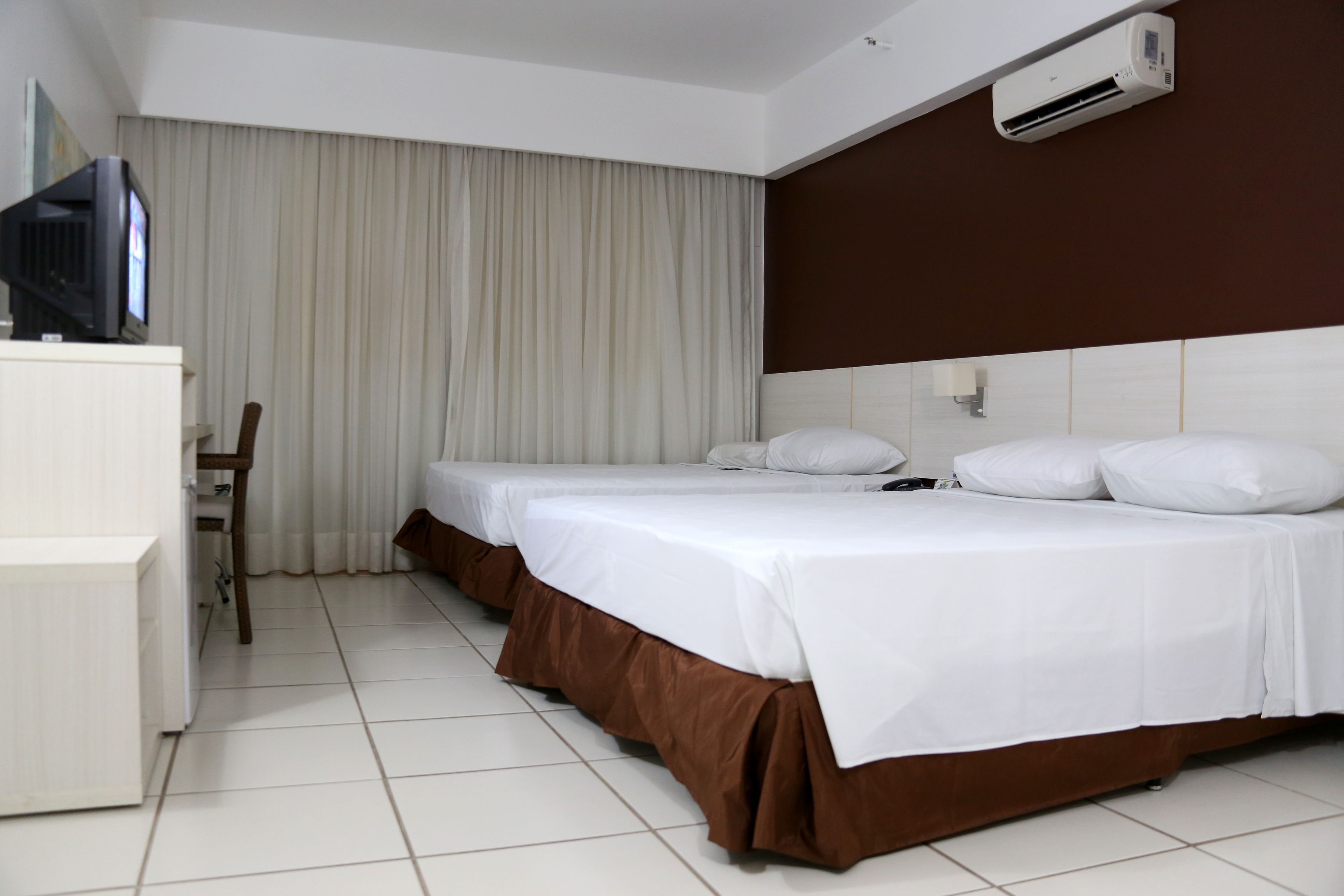 A hotel room with two beds, a desk, a television, and an air conditioning unit.