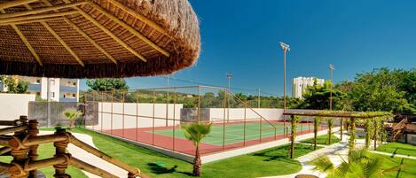 A tennis court with a net, surrounded by a fence, and a thatched roof structure.