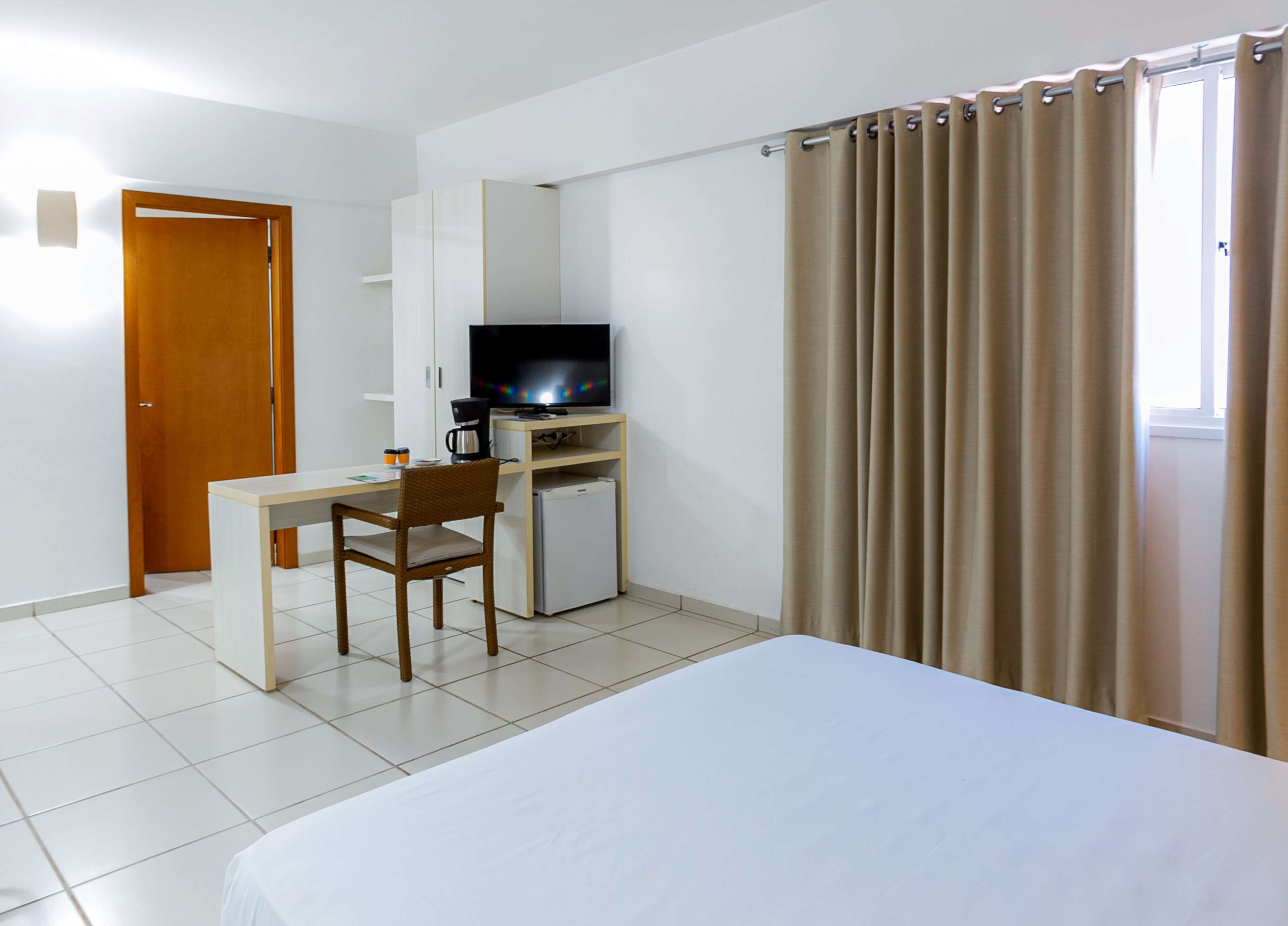 A hotel room with a bed, a desk with a television, a refrigerator, and a door.