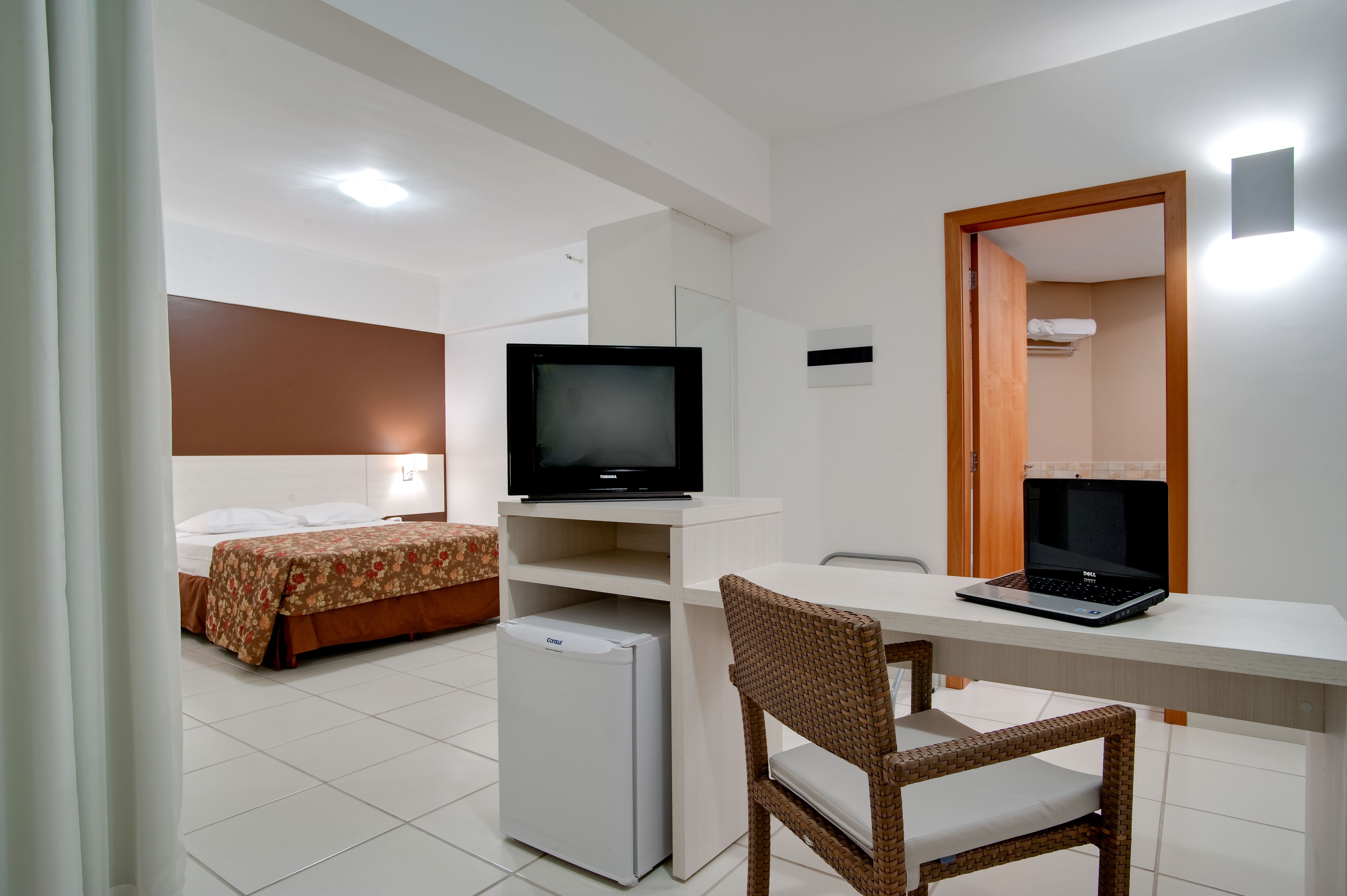 Una habitación de hotel con cama, escritorio, laptop, televisor y refrigerador.