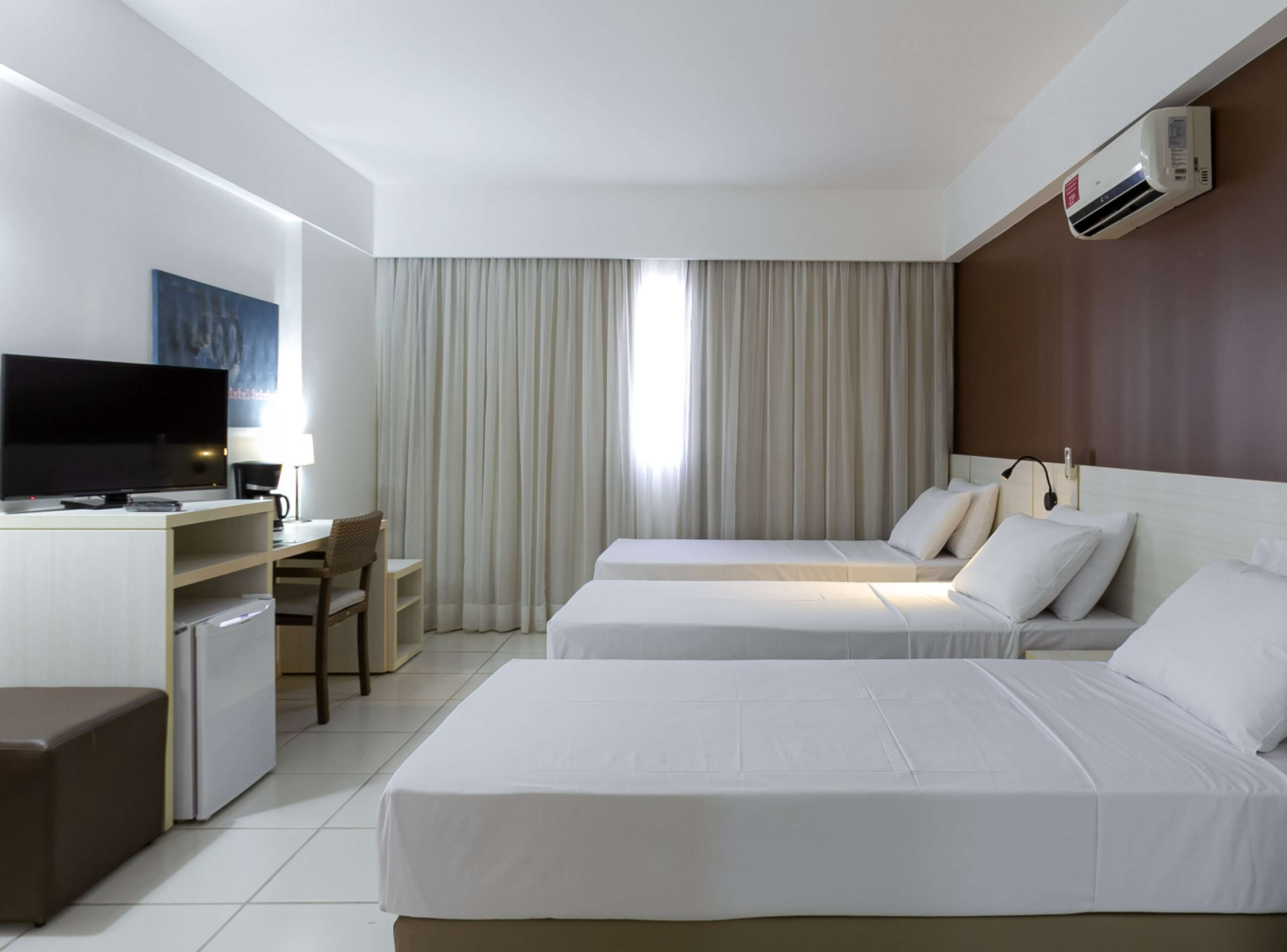 Ein Hotelzimmer mit zwei Betten, einem Schreibtisch mit Fernseher, einem Sessel, einer Lampe und einer Klimaanlage.