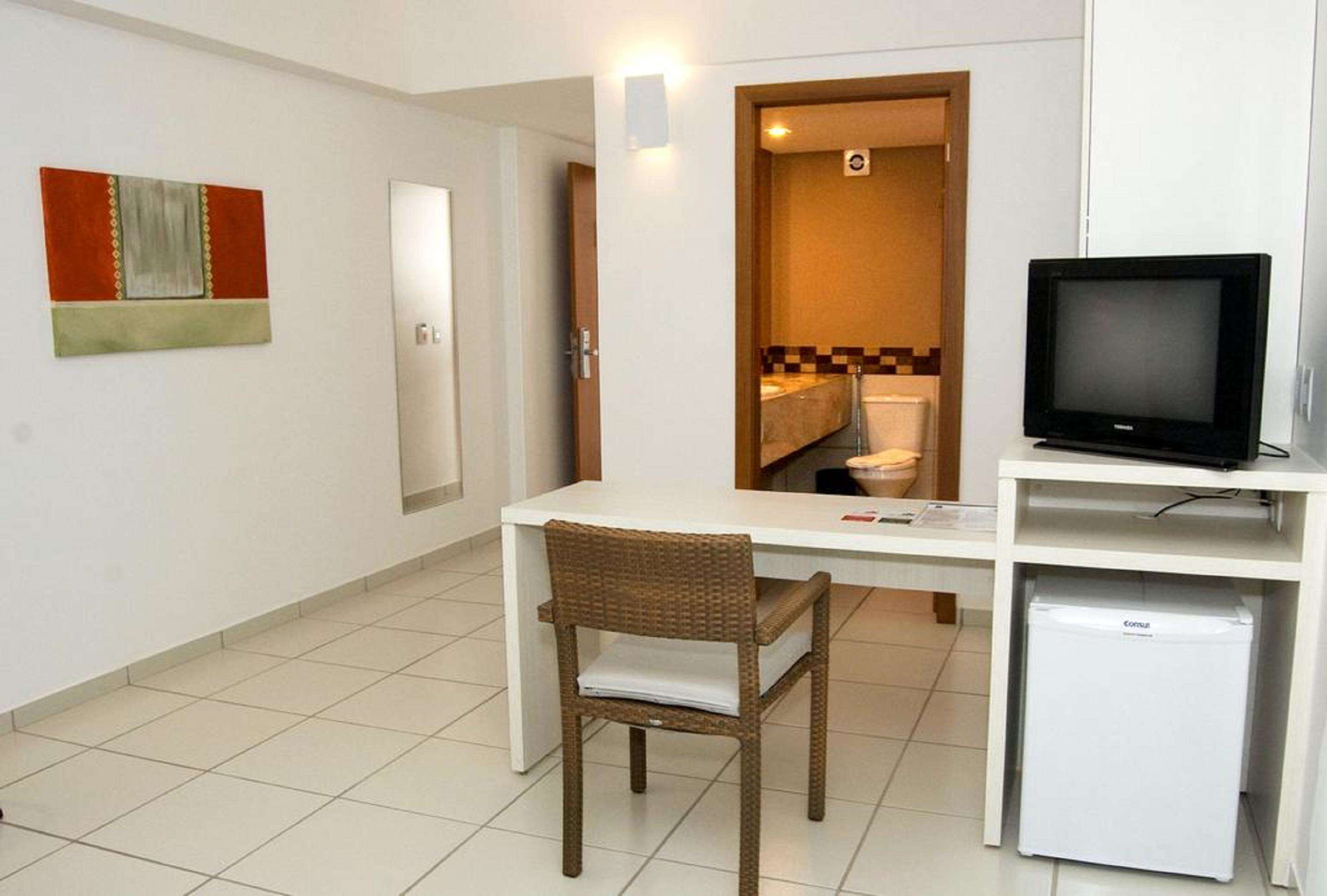 Une pièce de taille modeste, avec un bureau blanc, une chaise en osier, un petit réfrigérateur blanc et un téléviseur posé sur un meuble.