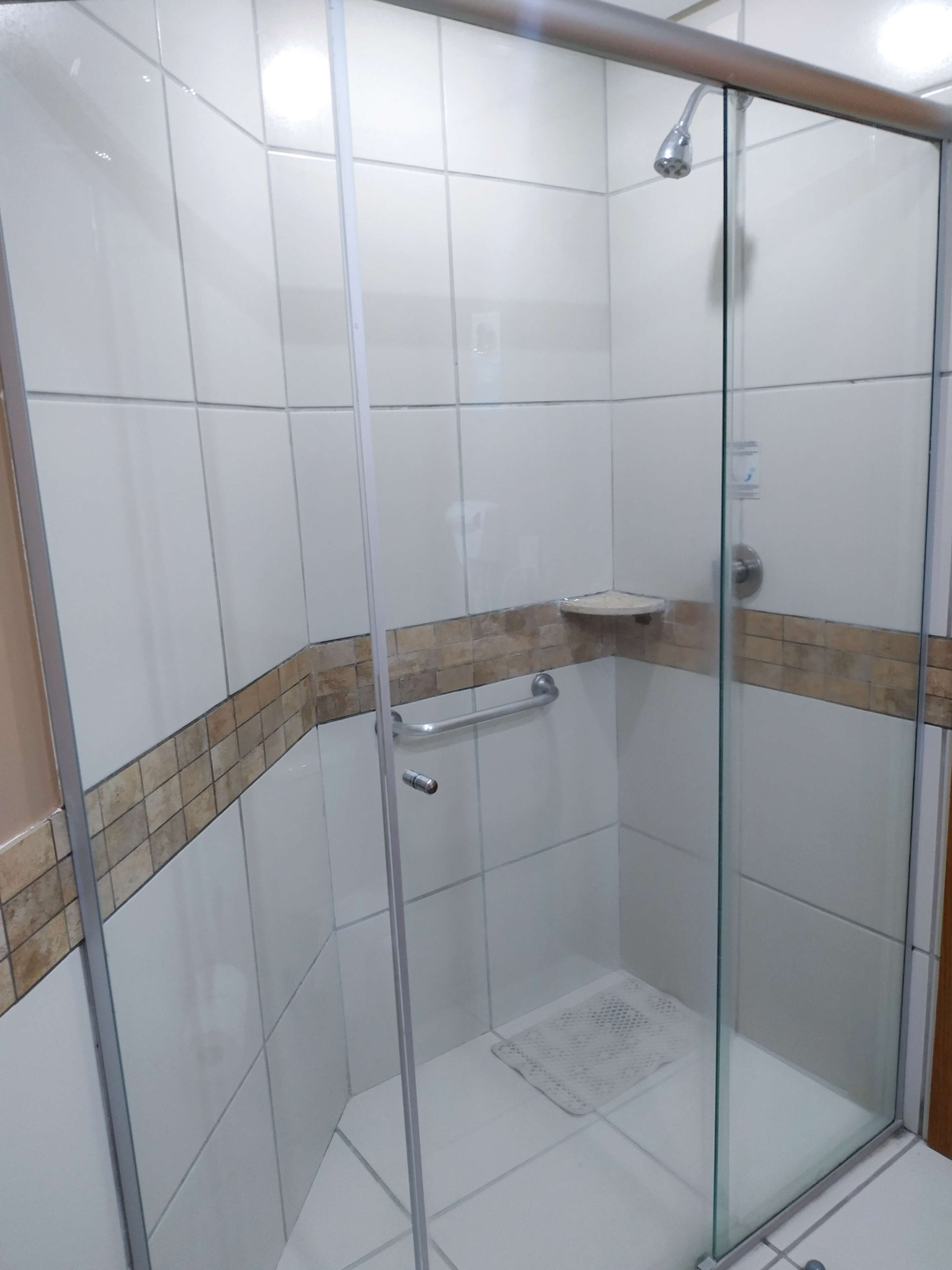 A glass-enclosed shower stall with a white tiled wall, a beige-tiled ledge, and a small white sink.