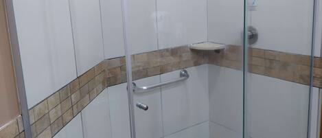 A glass-enclosed shower stall with a white tiled wall, a beige-tiled ledge, and a small white sink.
