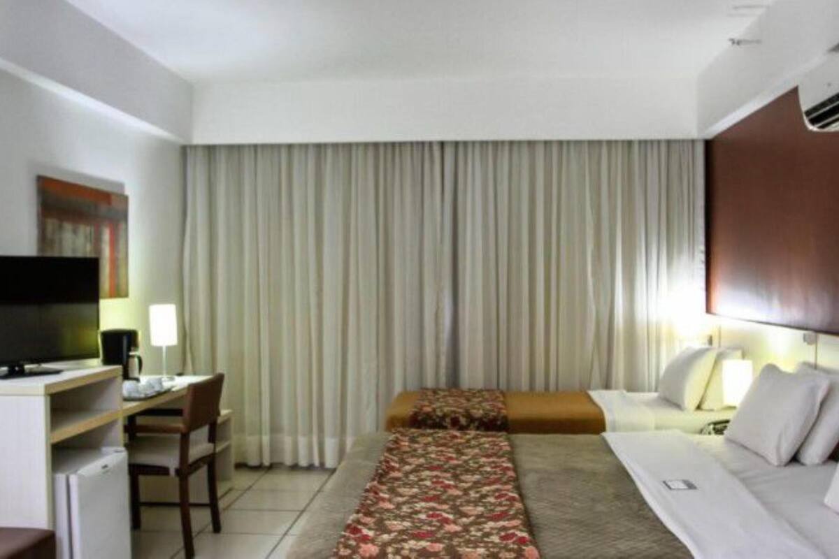 Una habitación de hotel con cama, escritorio con televisor, silla y ventana con cortinas.