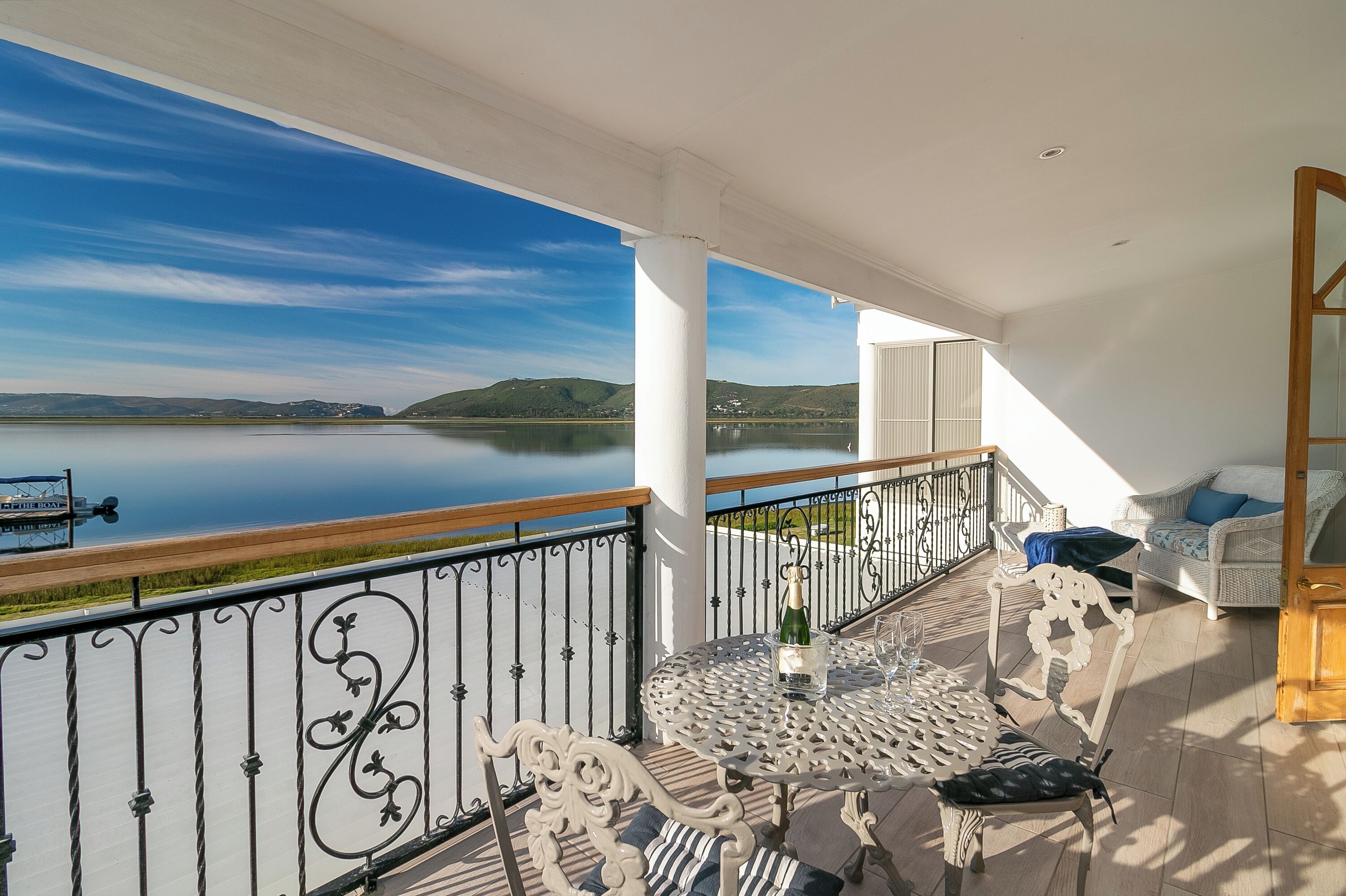A balcony with a view of a lake and hills, furnished with a table and chairs, and a sofa.