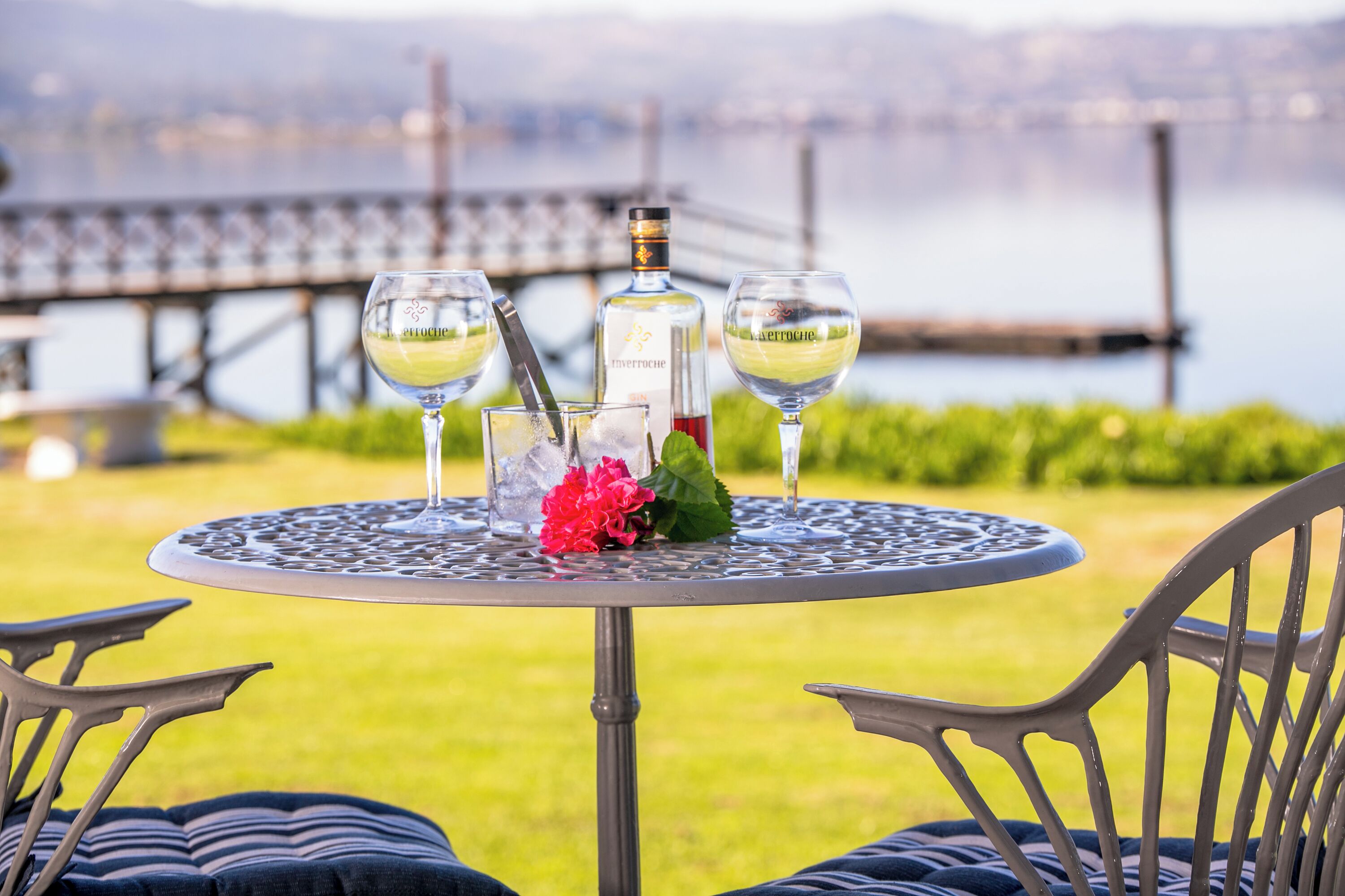A round metal table with three glasses of white wine, a bottle, and a red flower centerpiece.