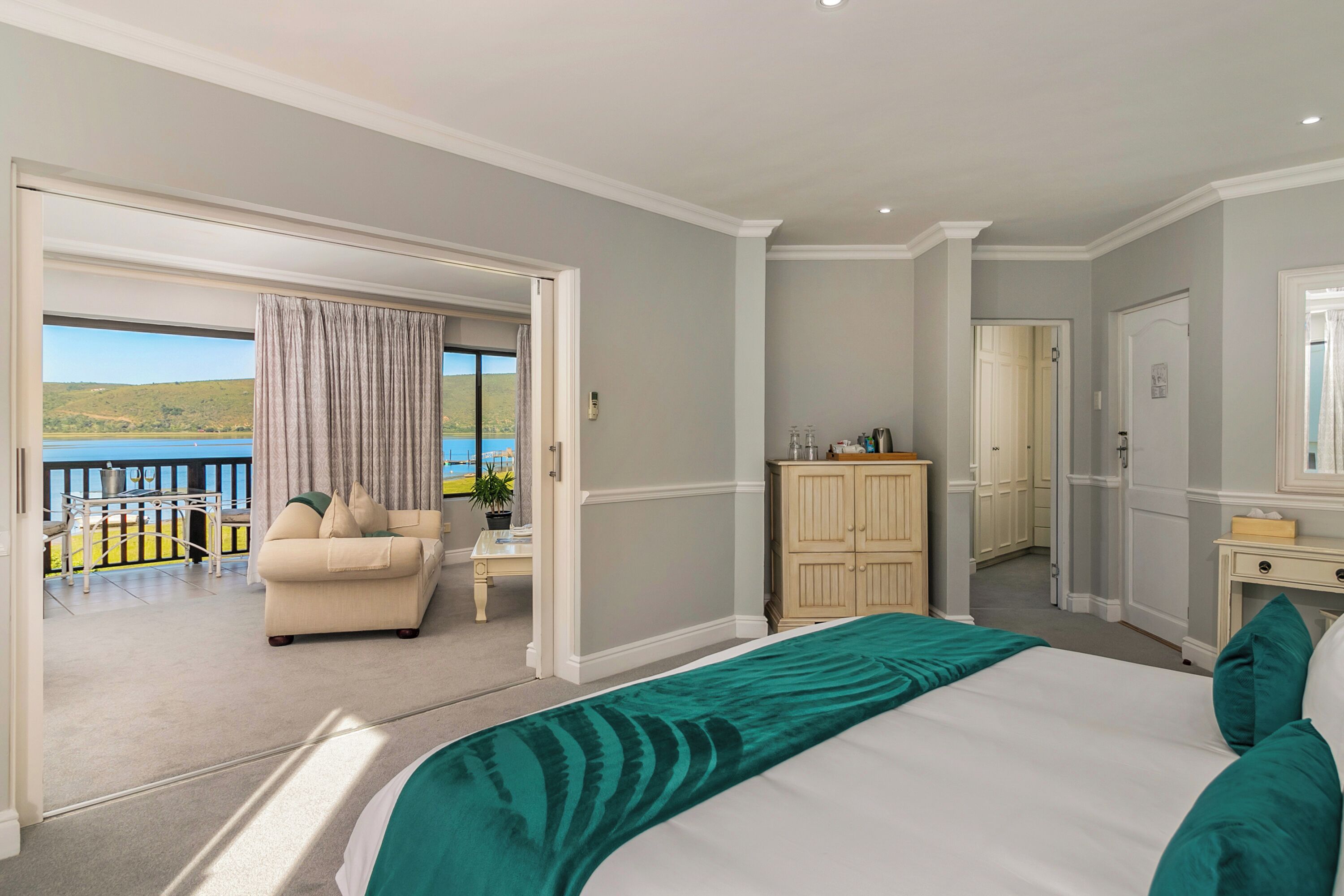 A bedroom with a large bed, a view of the outdoors, a seating area with a sofa and table, and a wardrobe.