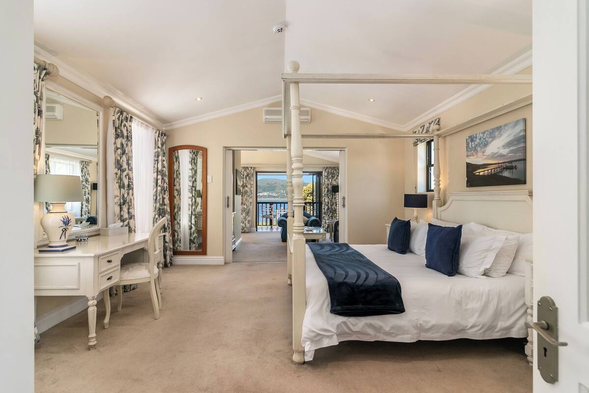 A bedroom with a four-poster bed, a desk with a chair, a mirror, and a view of the outdoors.