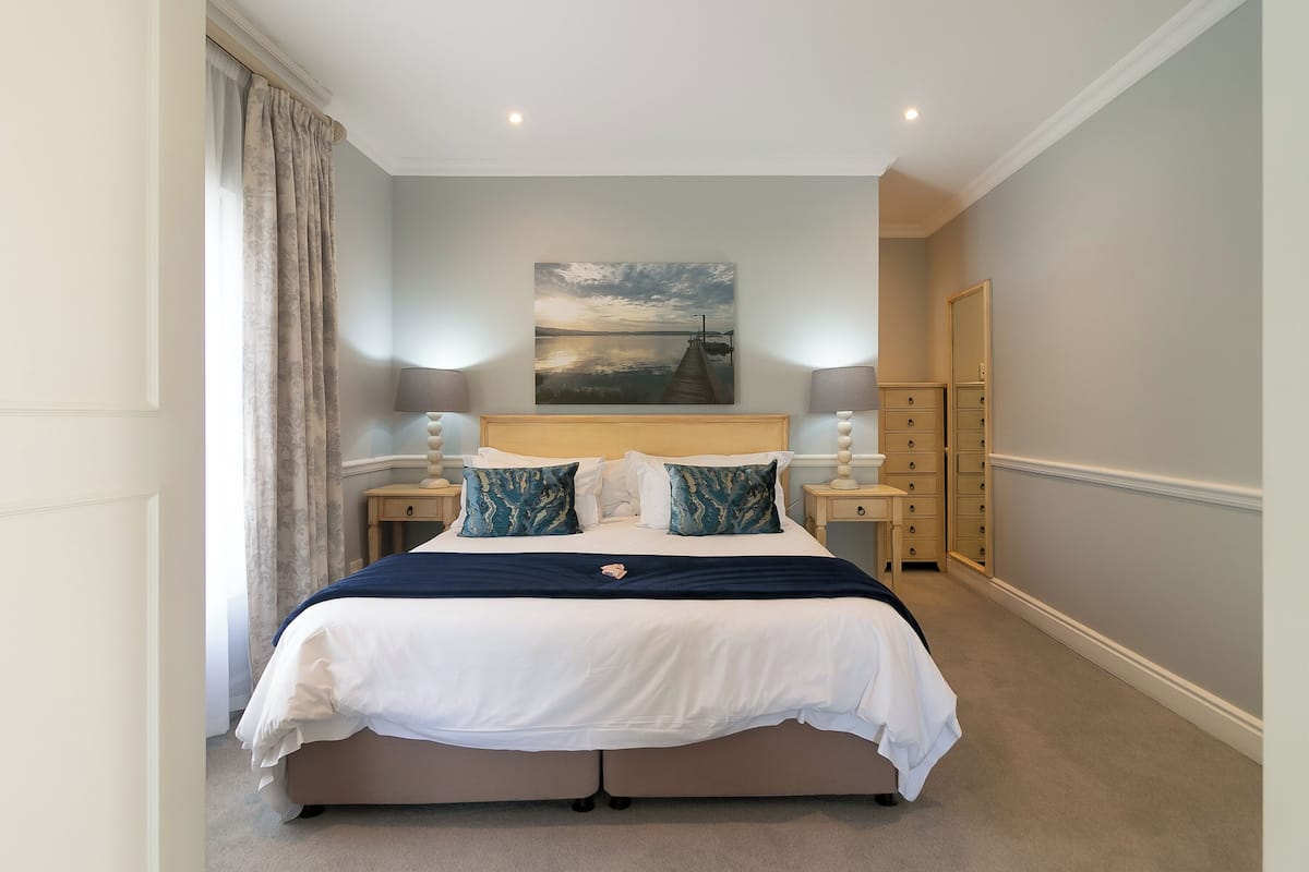 A neatly arranged bedroom with a bed, two bedside tables, a nightstand, a dresser, and a painting on the wall.