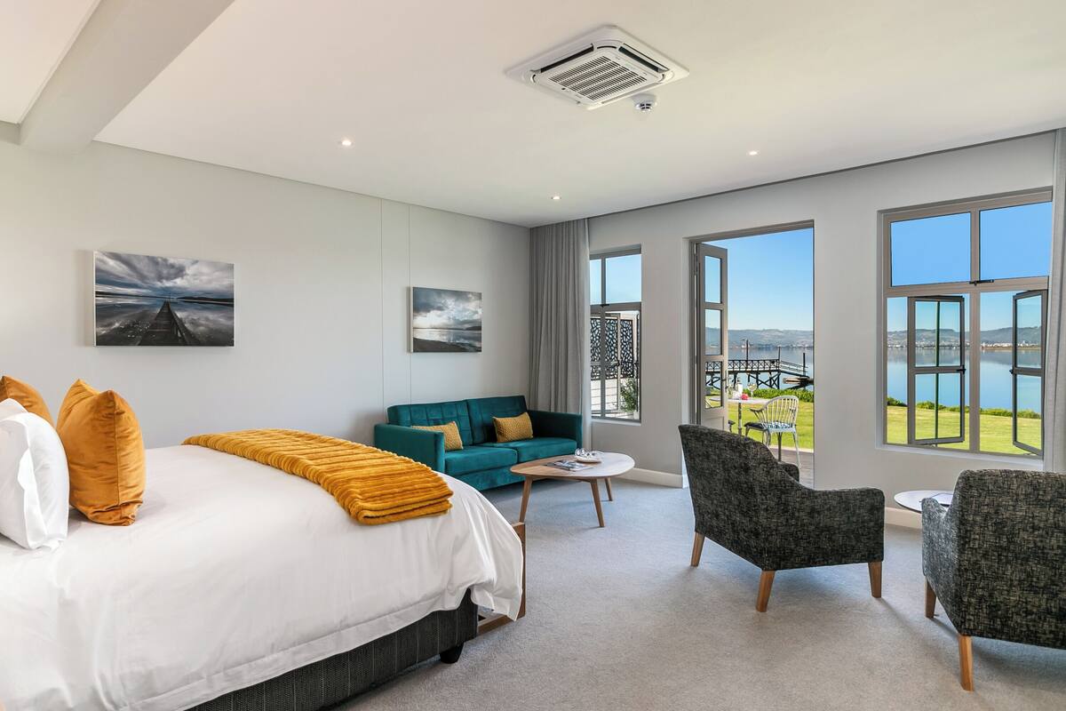 A modern hotel room with a large bed, a sofa, two armchairs, a small table, and large windows offering a view of the outdoors.
