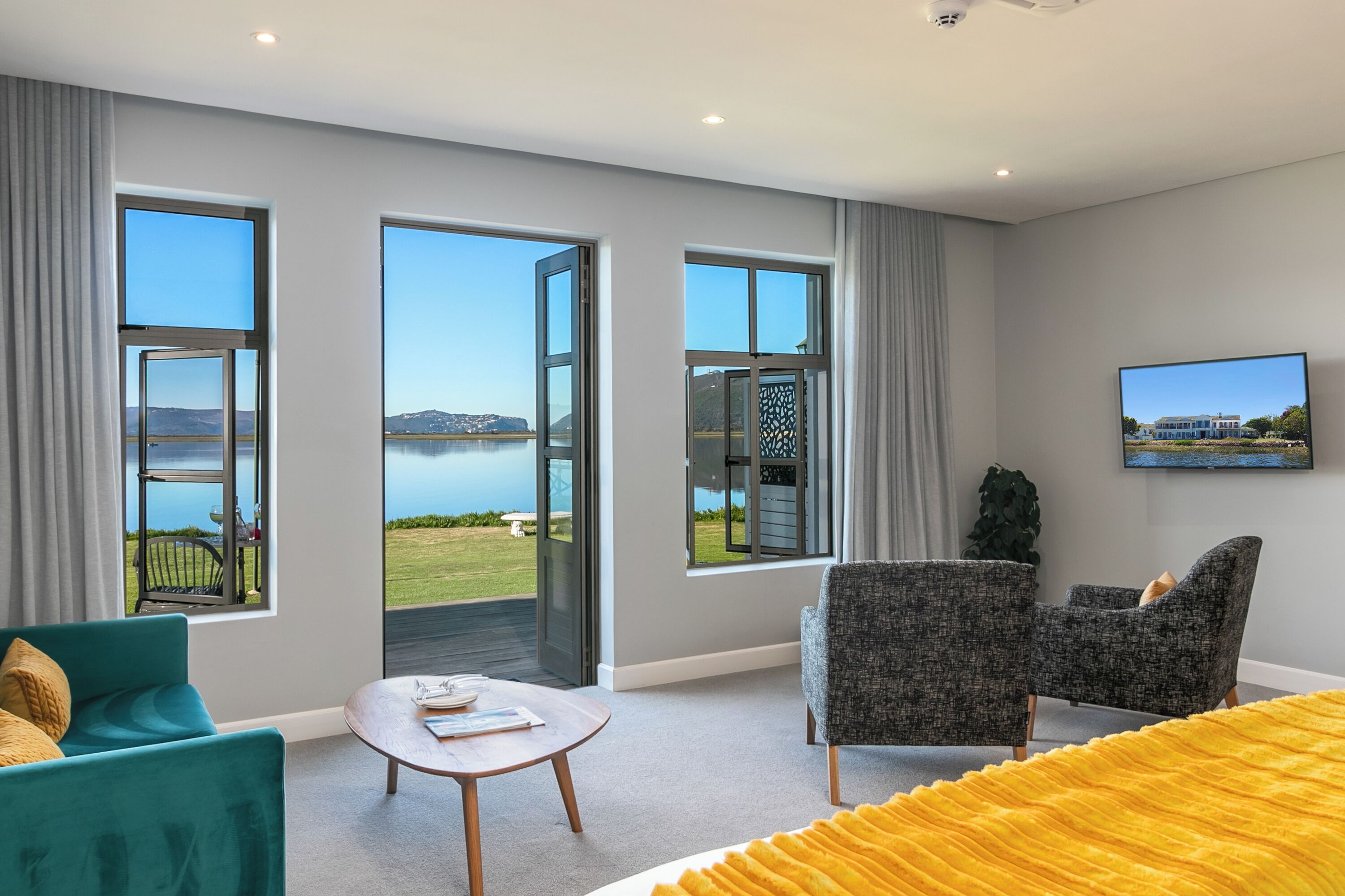 A modern hotel room with large windows offering a view of the lake and hills. There is a sofa, a chair, and a small table with a remote control.
