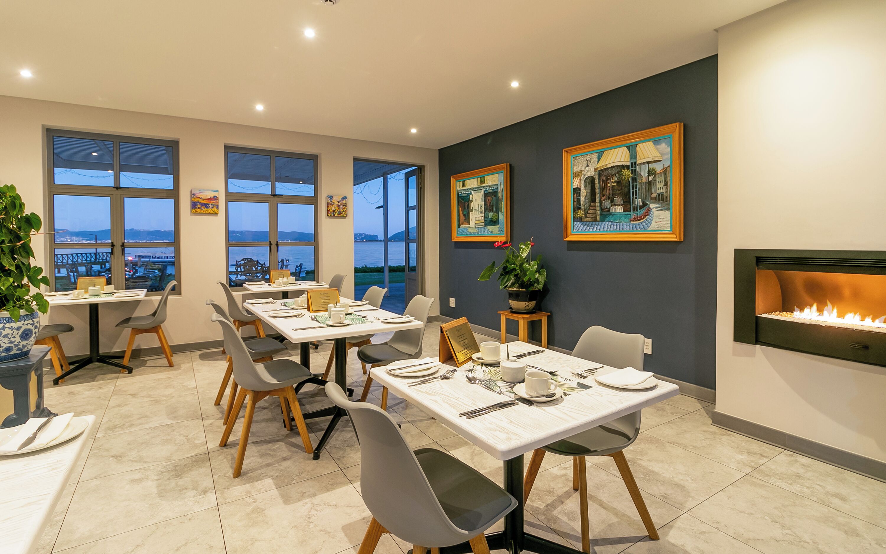 A dining area with tables set for two, large windows offering a view of the sea, and a fireplace on the wall.