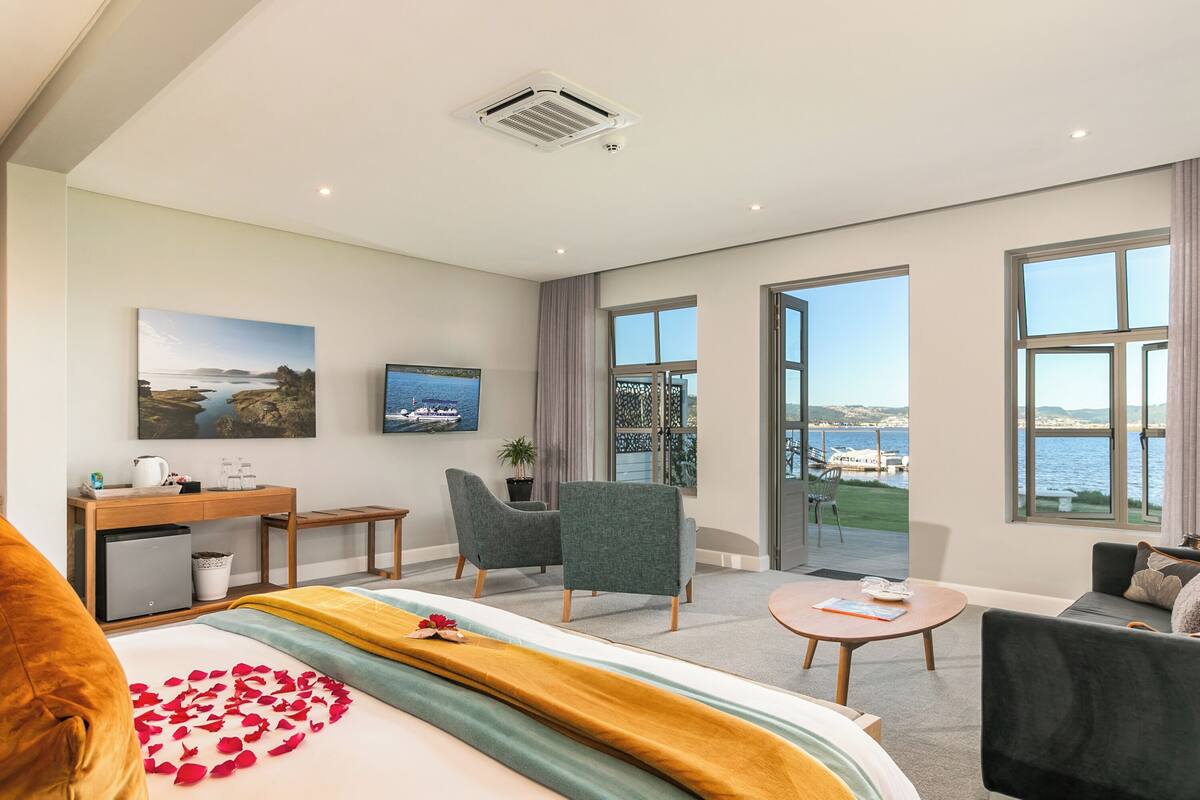 A modern hotel room with a bed, a desk, two chairs, a sofa, and large windows offering a view of the sea.