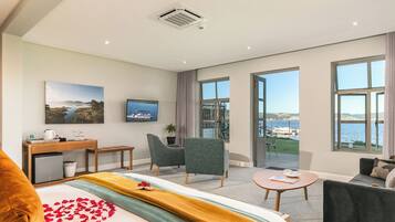A modern hotel room with a bed, a desk, two chairs, a sofa, and large windows offering a view of the sea.