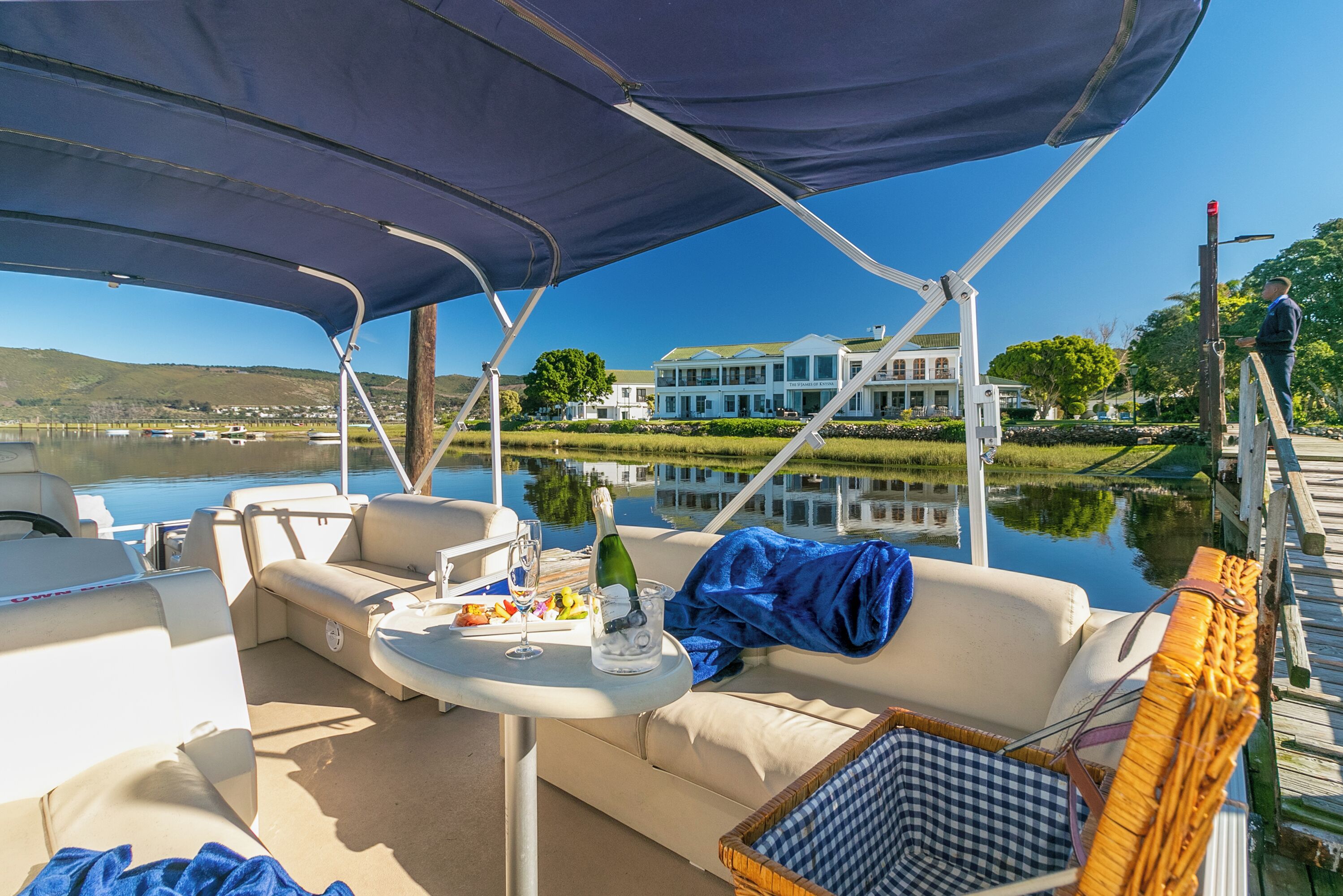A boat deck with a covered seating area, a table with a bottle and glasses, and a view of a large house across the water.