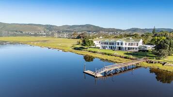 A large, white, two-story building with a green roof, situated by a calm lake, surrounded by grassy areas and hills in the background.