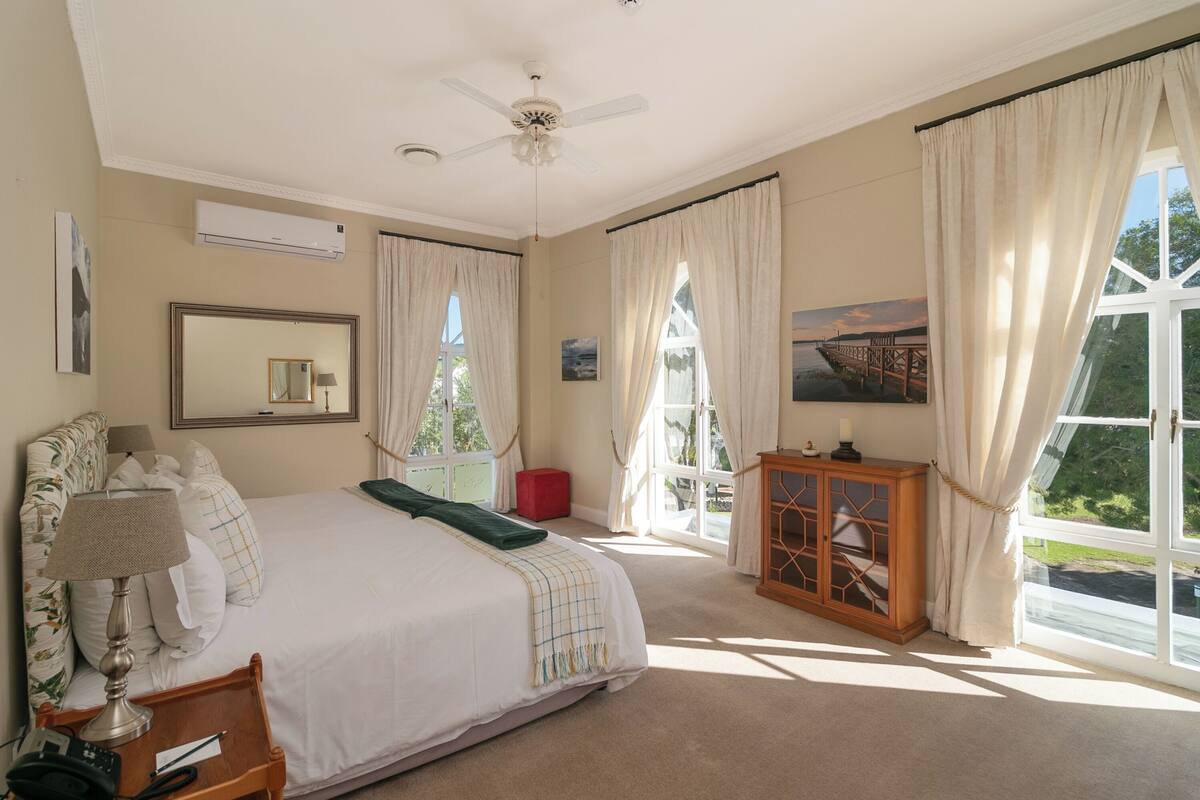 A bedroom with a large bed, a ceiling fan, an air conditioning unit, a wooden cabinet, and a window with curtains.