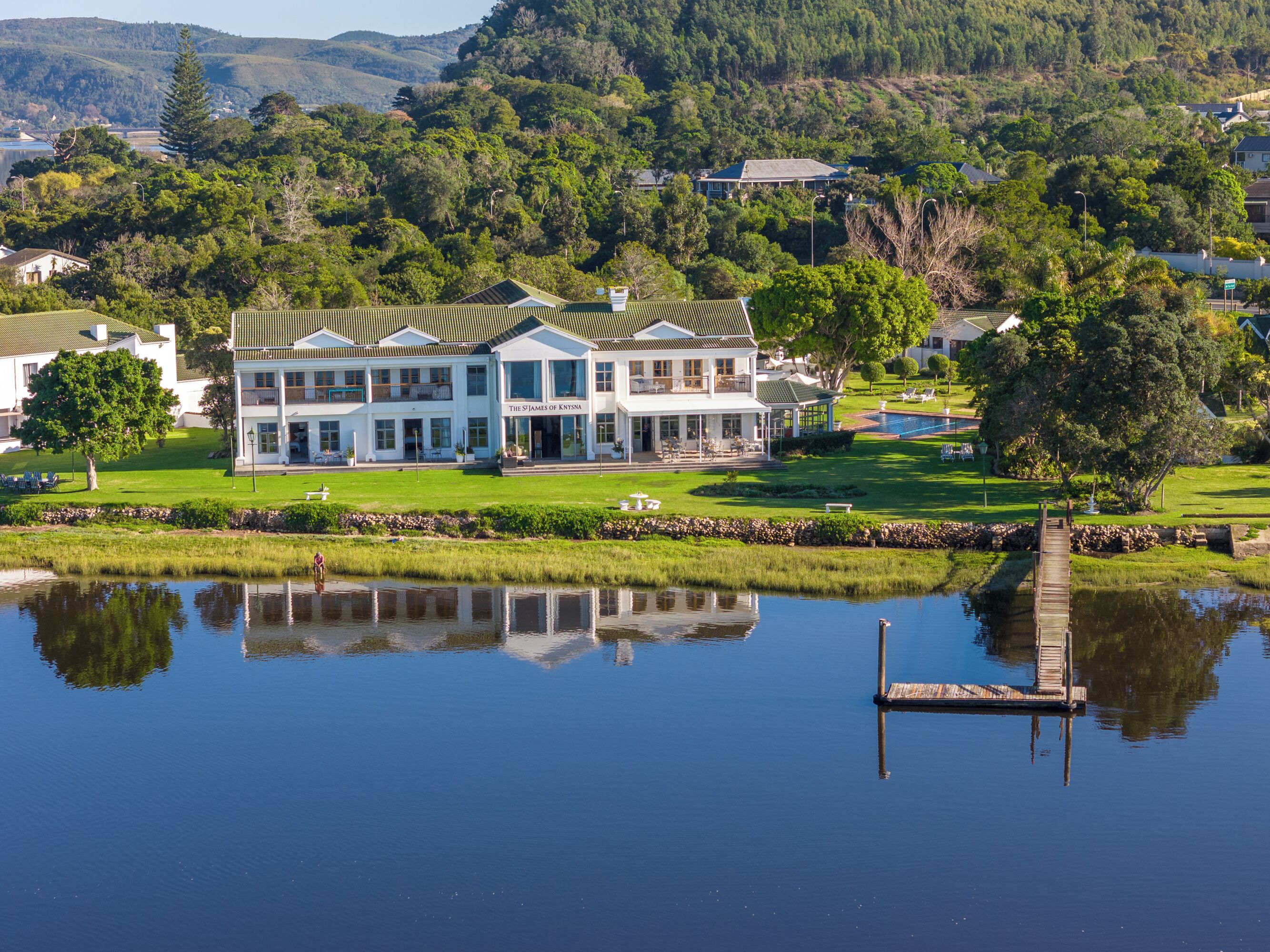 A large, two-story building with a green roof and a covered porch, surrounded by a well-maintained lawn and a body of water reflecting the structure.