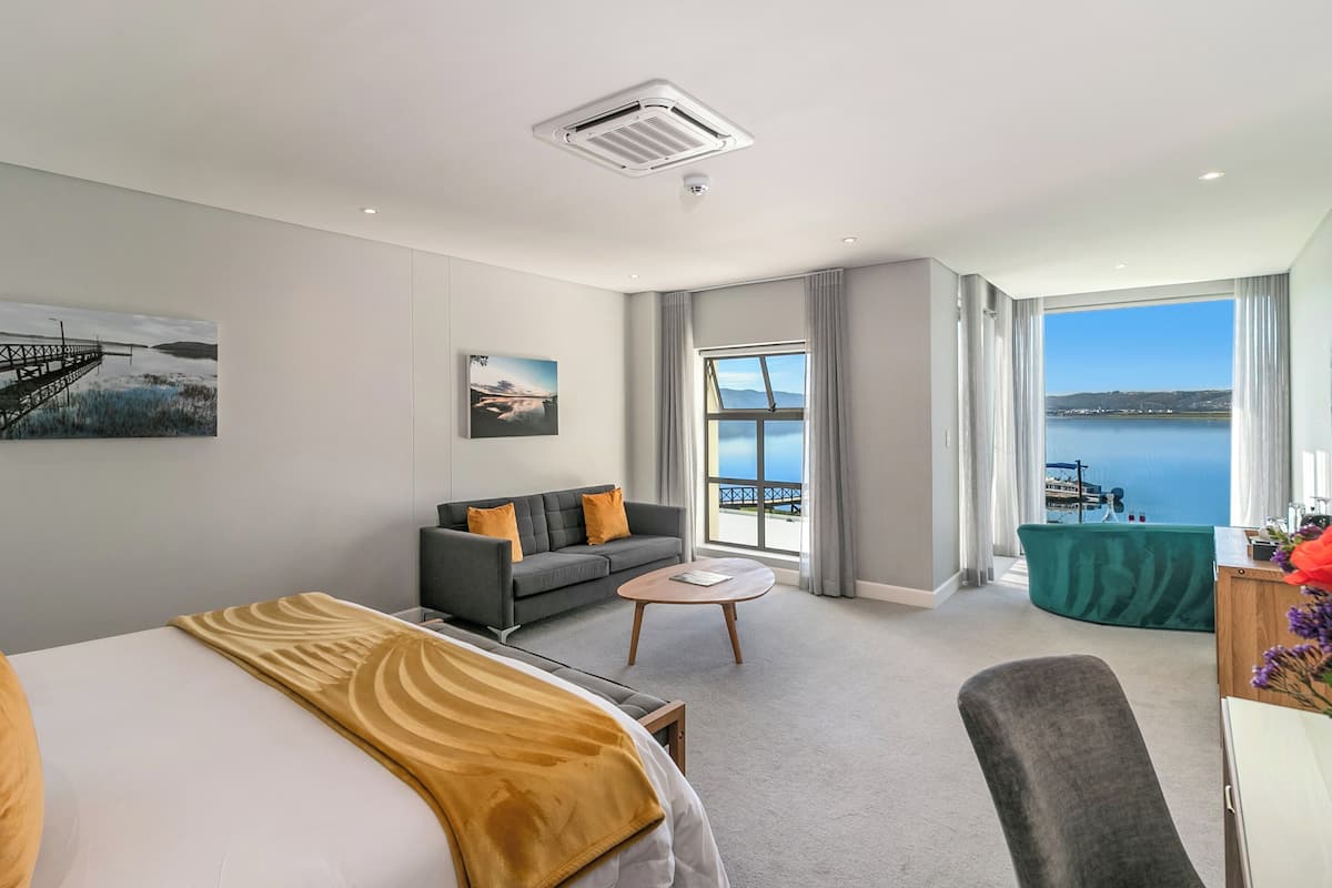 A modern hotel room with a bed, a sofa, a small coffee table, and a balcony with a view of the sea.