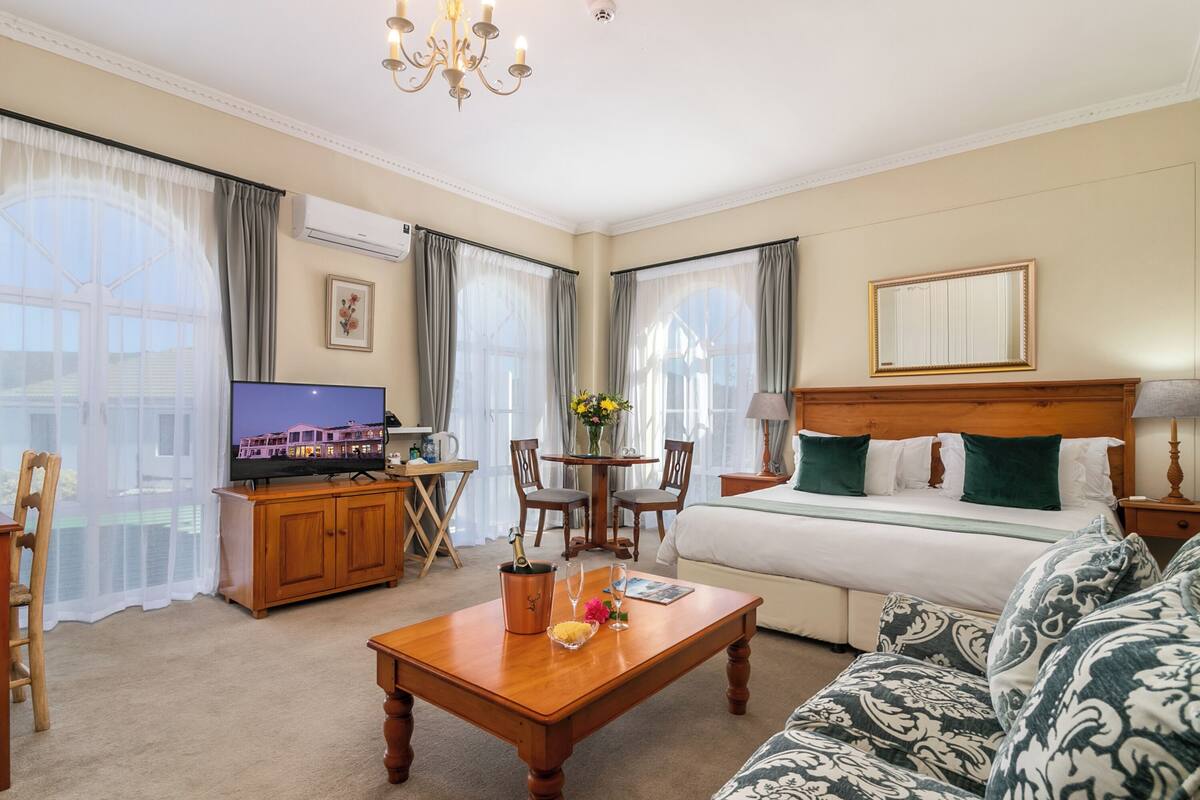 A hotel room with a bed, a sofa, a TV, a wooden coffee table, a desk with a chair, and a window with curtains.