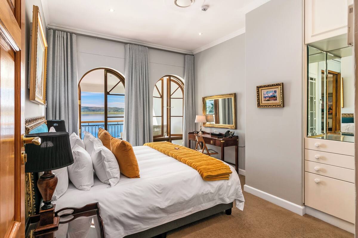A hotel room with a large bed, a desk, a chair, a mirror, and a view of the outdoors.