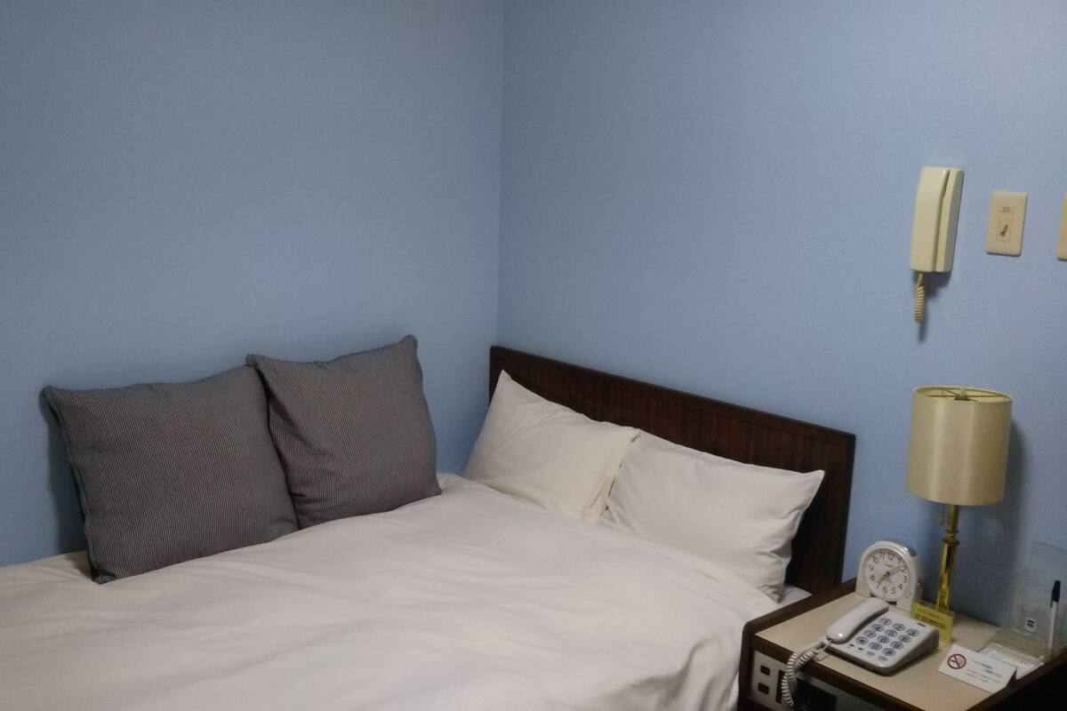 Une chambre d’hôtel avec un lit, deux oreillers, une table de chevet avec une lampe, un téléphone et une horloge.