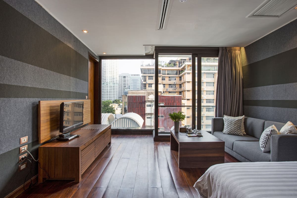 Habitación de hotel moderna con un escritorio de madera, un sofá, una cama y un gran ventanal con vistas a la ciudad.