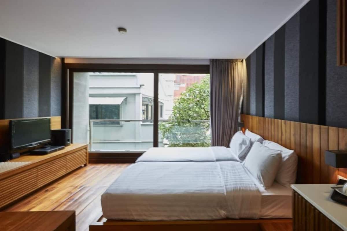 Habitación de hotel moderna con una cama grande, un televisor sobre un mueble de madera y vistas de la ciudad a través de un ventanal.