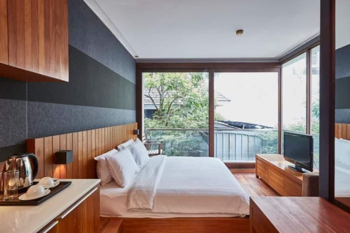 Una habitación de hotel moderna con una cama grande, televisión y balcón con vistas a árboles y un edificio.
