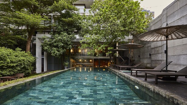 A swimming pool with a geometric tile pattern, surrounded by lounge chairs and umbrellas, with a building in the background. - LUXX Langsuan Hotel (Bangkok)