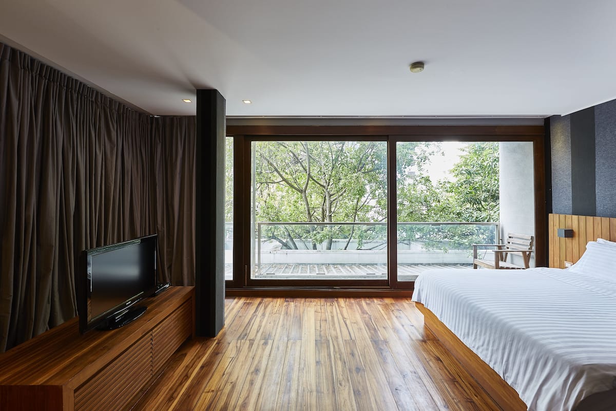 Habitación de hotel moderna con una cama grande, un televisor de pantalla plana sobre una consola de madera, un balcón con vistas a árboles y una puerta corredera de vidrio.