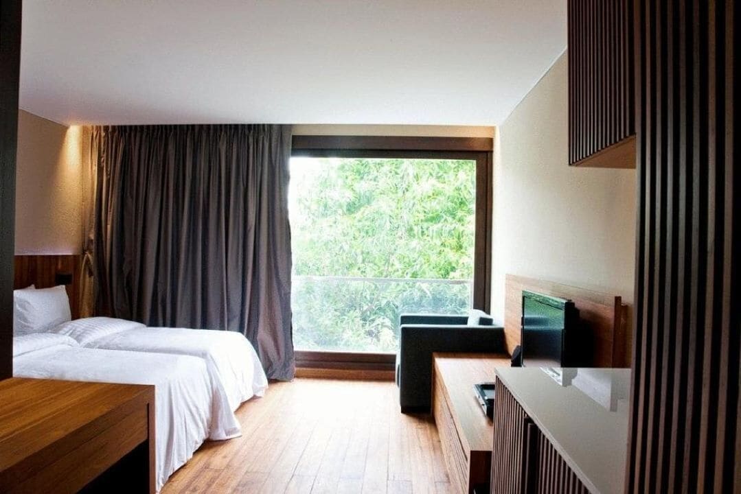 A hotel room with two beds, a wooden desk, a TV, and a balcony with a view of greenery.