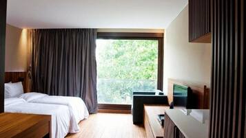 A hotel room with two beds, a wooden desk, a TV, and a balcony with a view of greenery.