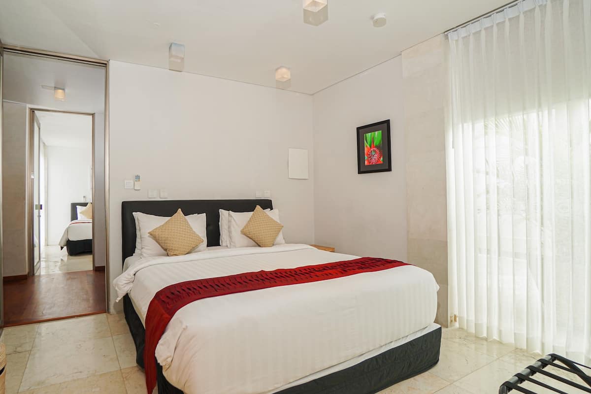 A hotel room with a large bed, a framed picture on the wall, and a view of a garden through sheer curtains.