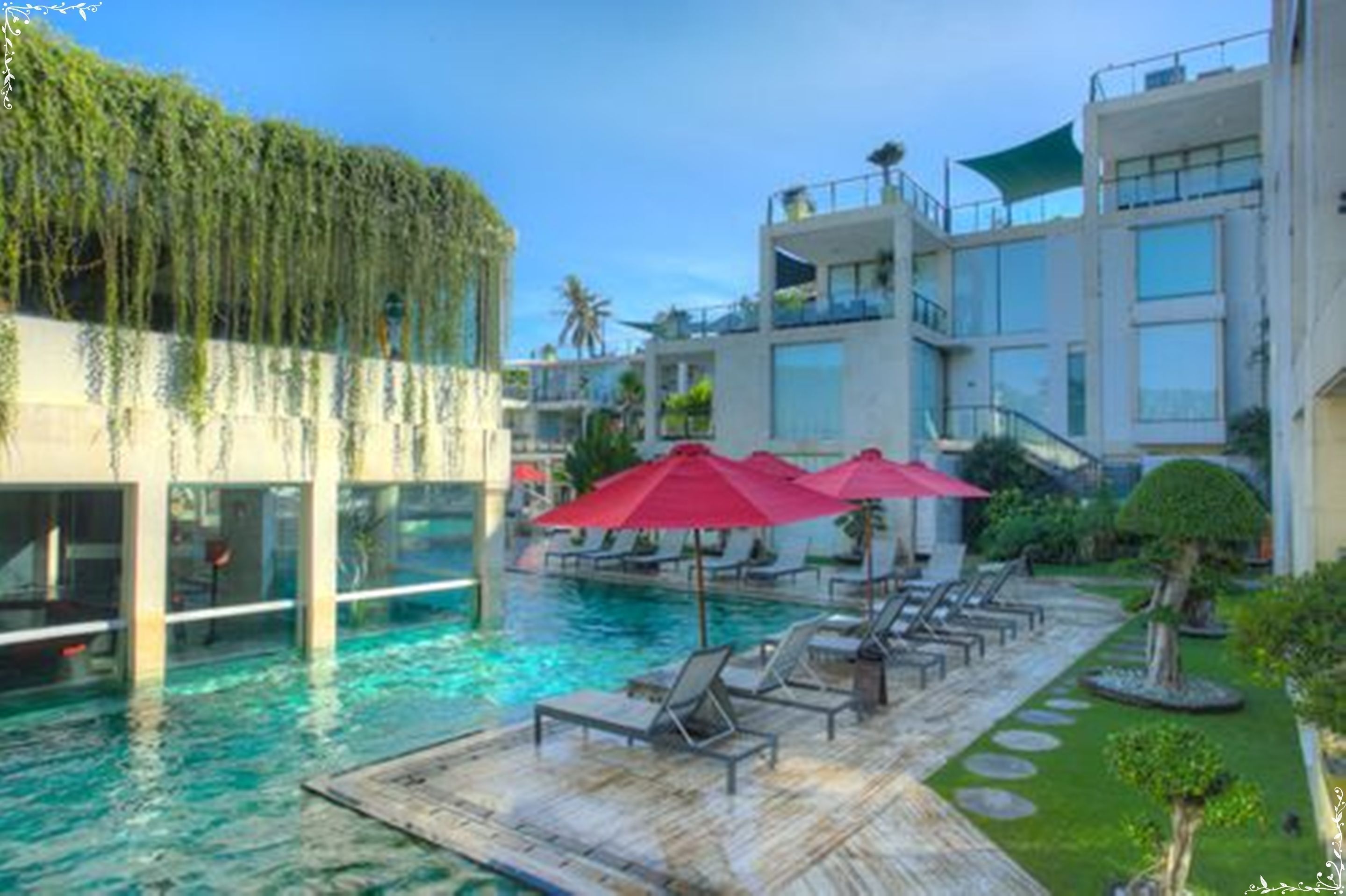 A pool area with lounge chairs and umbrellas, surrounded by modern buildings with balconies and a clear sky.