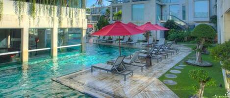 Zona de piscina con sillas de descanso y sombrillas, rodeada de edificios modernos con balcones y un cielo despejado.