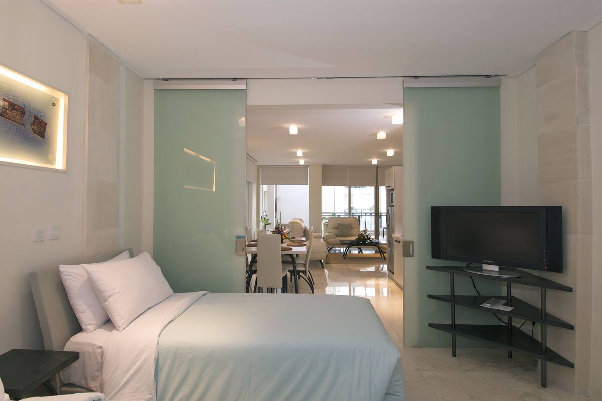 A modern bedroom with a bed, a TV on a stand, and a dining area in the background.