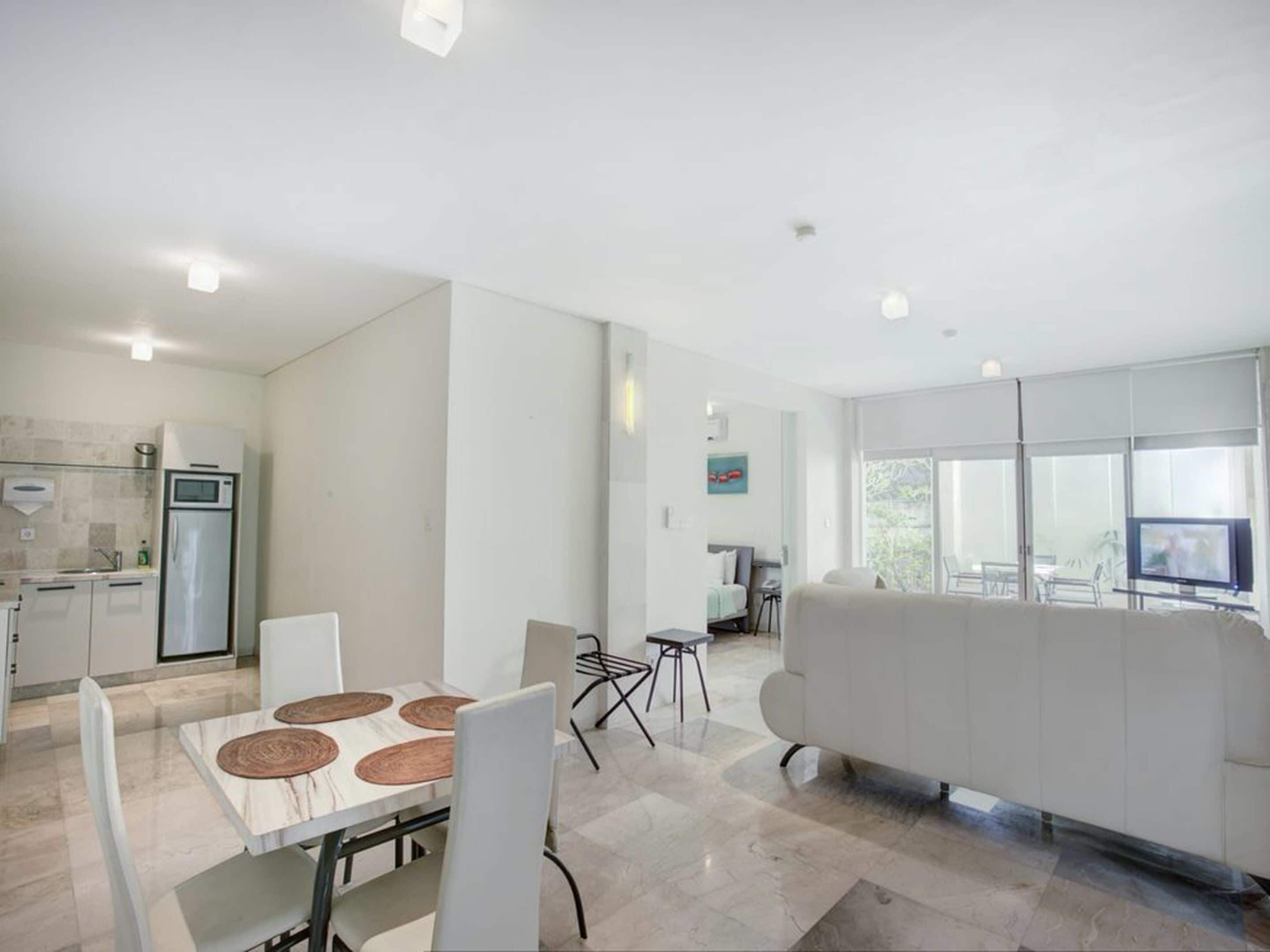 Een moderne keuken met een koelkast, magnetron en spoelbak. Er is een eethoek met een tafel en stoelen. In de woonkamer staat een witte bank, en een televisie is aan de muur bevestigd.